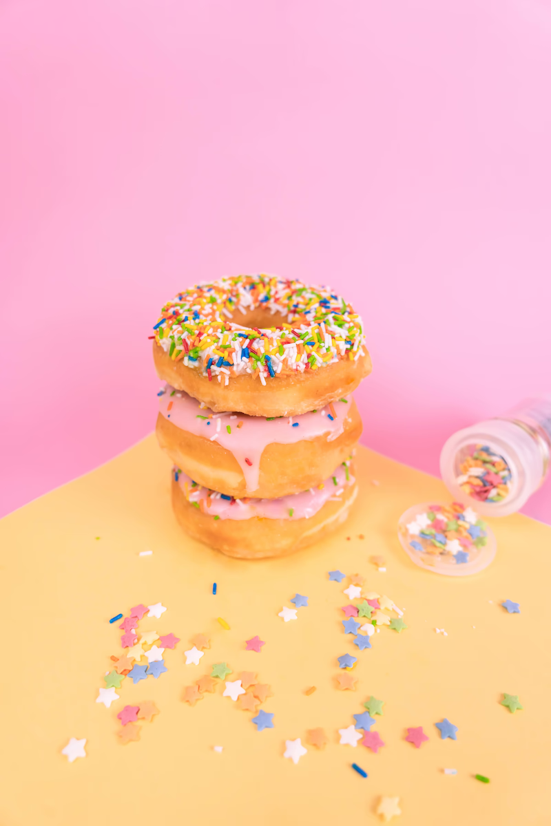 Three stacked glazed donuts with sprinks on a pink and yellow background.